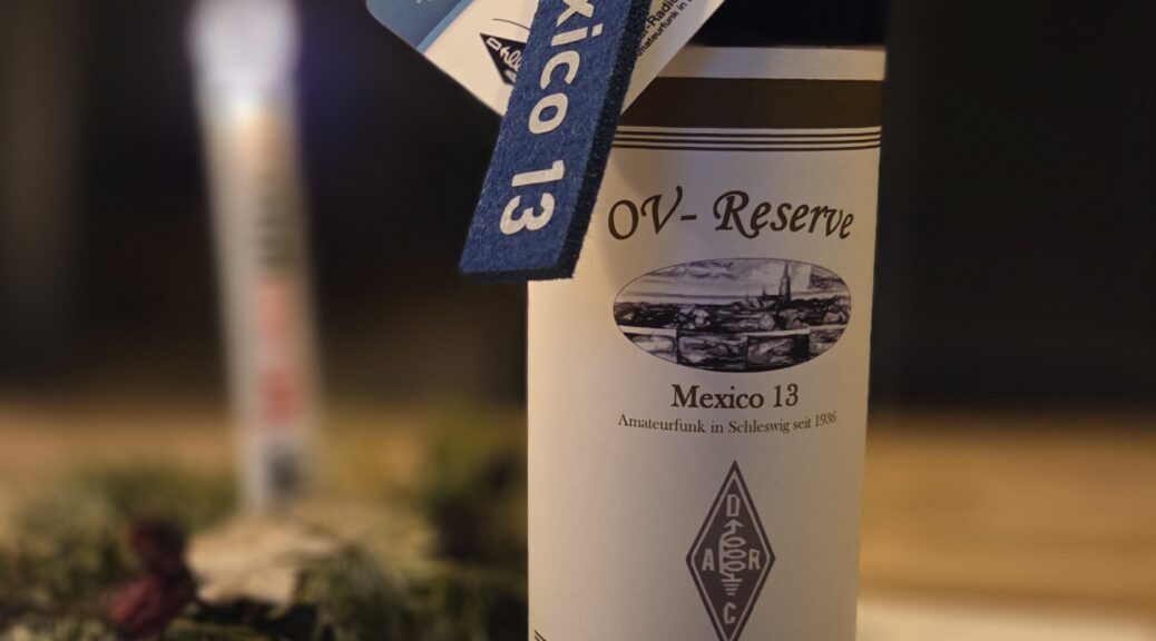 Rotweinflasche mit OV-Wein des DARC-Ortsverbands M13 Schleswig. Daran hängt ein Mexico 13-Schlüsselanhänger. Weiterhin eine Urkunde.